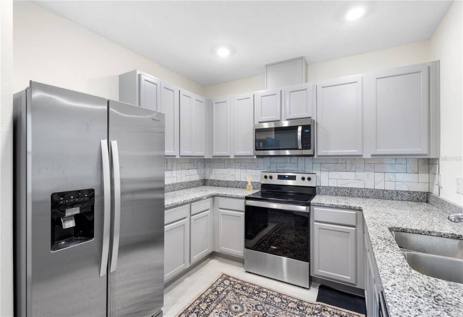 Kitchen with Living Room and Primary Suite Behind. Ahead lies the Foyer with Hallway leading to Garage/Laundry Room, Dining Area, and Hallway leading to Secondary Bedrooms and Bath.